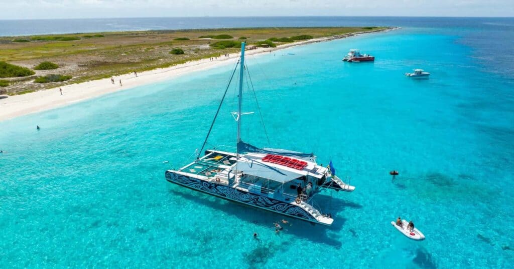 A colorful catamaran floats on clear turquoise water near a sandy beach with green vegetation, showcasing featured products as people swim, snorkel, and relax on boats under a sunny sky. Other boats are anchored nearby along the shoreline.